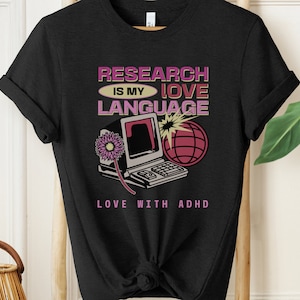 Op de afbeelding: Zwart t-shirt met een retro design. De afbeelding bevat de tekst "RESEARCH IS MY LOVE LANGUAGE" boven een computer, wereldbol en bloem illustratie. De tekst "LOVE WITH ADHD" staat eronder.