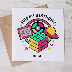 May include: A white card with a colourful birthday greeting. The greeting says "Happy Birthday" in a rainbow arc with a retro 80s theme. The card features a Rubik's Cube, a toy Etch A Sketch, a flying saucer, and a spinning wheel. The name "Angie" is printed at the bottom of the card.