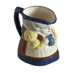 May include: A ceramic character jug shaped like a person wearing a brown dress with yellow sleeves and a white apron. The figure holds a small blue pitcher. The jug features a dark blue handle, a blue rim, and a blue base, with painted facial features.