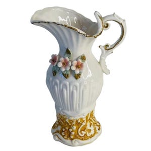 May include: White ceramic pitcher with a gold rim and handle. The pitcher features embossed vertical lines and is decorated with pink and white flowers and green leaves. The base has a gold floral design.