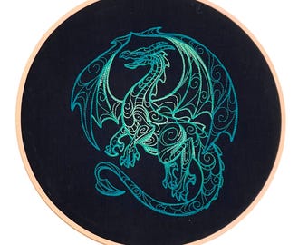 Puede incluir: Un bastidor de bordado circular de madera que contiene tela negra con un diseño de dragón detallado cosido en hilo verde y azul turquesa vibrante. El dragón se representa en un patrón de remolinos con las alas extendidas y una cola larga.