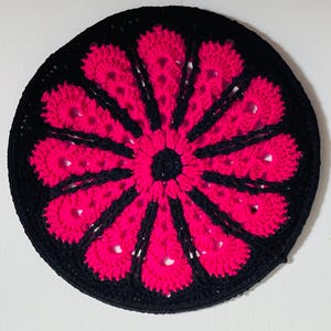 May include: A round crocheted coaster featuring a black and pink floral design. The centre of the flower is black and the petals are pink.