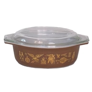 May include: A brown casserole dish with a clear glass lid. The dish is decorated with gold-colored illustrations of various kitchen-related items, including a rooster, eagle, and wheat. The dish has handles on either side.
