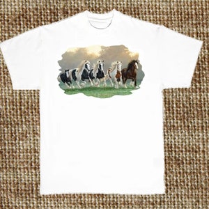 Pode incluir: Camiseta branca com uma imagem colorida de cinco cavalos correndo por um campo. Os cavalos são pretos, brancos, marrons e manchados.