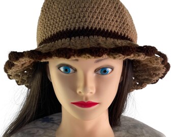 May include: A crocheted hat in shades of brown. The hat features a wide brim with a scalloped edge and a dark brown band around the crown. The hat is displayed on a mannequin head with blue eyes and red lipstick.