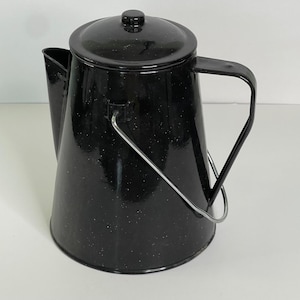 May include: A black enamelware coffee pot with a lid and a wire handle. The pot has a speckled pattern and a spout for pouring. The handle is made of metal. The pot is designed for camping or outdoor use.