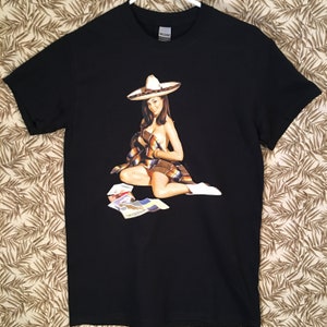 Könnte beinhalten: Schwarzes T-Shirt mit einem Aufdruck einer Frau, die einen Sombrero und eine Decke trägt und auf einem Stapel Papier sitzt.