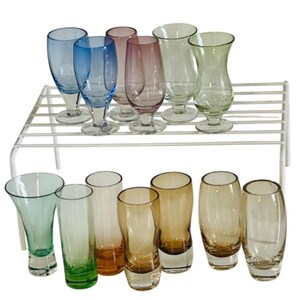 Puede incluir: Una colección de trece vasos de diferentes colores, incluyendo azul, rosa, verde y ámbar. Los vasos presentan formas variadas, algunos con bordes acampanados y otros con lados rectos, todos colocados sobre un estante de alambre de color blanco.