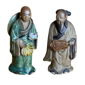 May include: Two ceramic figurines depicting standing figures in traditional robes. The left figure wears a green robe, holds a small white bowl and a yellow fan. The right figure wears a tan robe with dark accents and holds a brown fan.