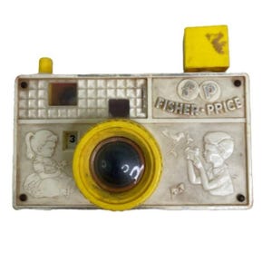 May include: Vintage Fisher-Price toy camera in off-white and yellow. The camera features a yellow lens, a yellow flash, and embossed images of children and the Fisher-Price logo. The number "3" is visible on the camera.