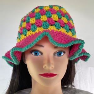 May include: A colorful crochet hat with a wide brim. The hat features a pattern of pink, yellow, and teal squares. The brim is pink with a teal trim. This handmade hat is a stylish accessory.