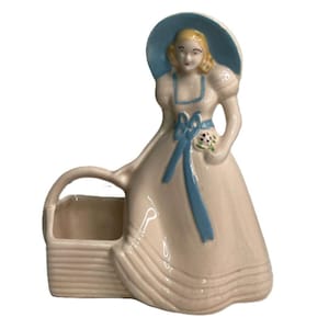 May include: A ceramic figurine planter shaped like a person wearing a wide-brimmed blue hat and a pale pink dress with a blue ribbon sash. The figure holds a small bouquet of flowers and stands beside a rectangular basket-weave container.