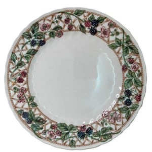 May include: White ceramic plate with a decorative border featuring a lattice design, green leaves, and clusters of red and dark blue berries. The plate has a wide rim with a brown rope-like edge.