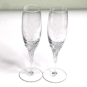 May include: Two clear champagne flutes with etched floral patterns. The stems have a textured, twisted design, and the bases are round. The flutes are presented on a white surface, suitable for a celebration or event.