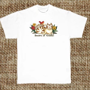 May include: White t-shirt featuring a cartoon illustration of a cat with brown and white stripes, a red bird, and flowers. The text "Beware of Furballs!" is printed below the image.