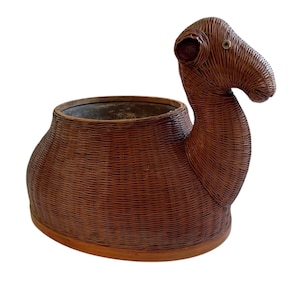 Puede incluir: Una jardinera con forma de camello de mimbre marrón. La jardinera tiene un recipiente grande y abierto para plantas, con la cabeza y el cuello del camello formando un elemento decorativo. El ojo del camello es un pequeño detalle redondo de color dorado.