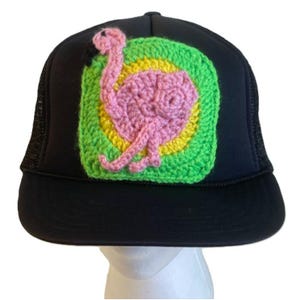 May include: Black trucker hat featuring a crocheted flamingo. The flamingo is pink with a black beak, set on a green, yellow, and white background. The hat has a mesh back and a curved peak.