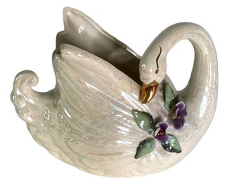 May include: A pearlescent ceramic swan-shaped planter with a gold beak and purple flower accents. The swan has a curved neck and a wide opening for plants or decor. The planter is a decorative item.