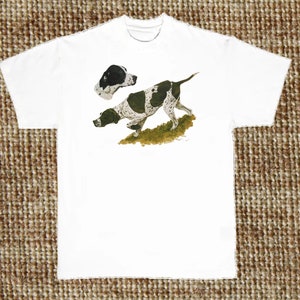 Op de afbeelding: Wit T-shirt met een zwart-witte illustratie van een jagende hond die wijst. De hond staat in een rennende houding met zijn hoofd naar de zijkant gedraaid. De illustratie bevindt zich op de voorkant van het shirt.