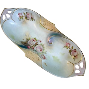 May include: An oval porcelain dish with a pale blue and white base, adorned with pink roses and gold detailing. The edges feature decorative cutouts. A vintage decorative piece, likely from the early 20th century.