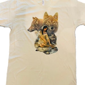 Peut inclure: Un t-shirt beige avec un imprimé graphique d'une femme amérindienne assise avec trois loups. La femme porte une robe à perles et tient un filet. Les loups sont bruns et gris.