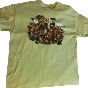 May include: A light green t-shirt with a printed image of a brown horse and a brown foal standing in a field of flowers.