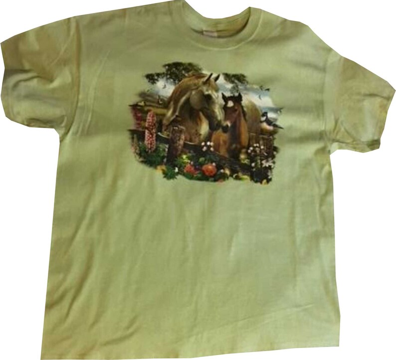 May include: A light green t-shirt with a printed image of a brown horse and a brown foal standing in a field of flowers.