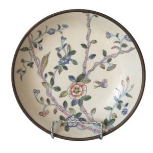 May include: A round, cream-colored decorative plate with a brown rim. The plate features a floral design with pink, blue, and green accents. The design includes flowers, leaves, and branches. The plate is displayed on a silver metal stand.