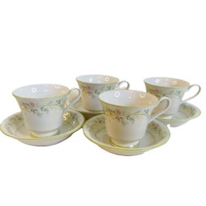 May include: Four white teacups and saucers with a floral design and a green trim. The cups have a curved handle and sit on matching saucers. The floral pattern includes pink and yellow flowers.