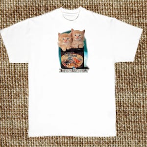 May include: White t-shirt with a graphic of two orange kittens peeking out of a vintage tin can labeled "Whiskers Double Trouble".