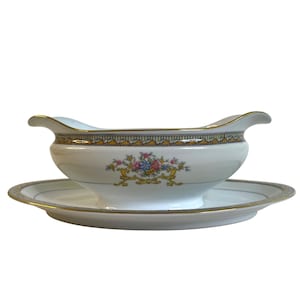 Puede incluir: Salsera de porcelana blanca con su platillo a juego. El borde es dorado y tiene una banda decorativa con un patrón geométrico. Un diseño floral con flores rosas, azules y amarillas adorna el frente.