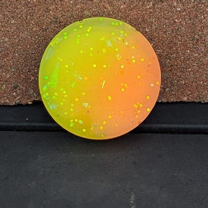 May include: A round, resin object with a gradient of yellow to orange. The object is speckled with small, glitter-like particles. The surface appears smooth and glossy, reflecting light. The item is a decorative element.