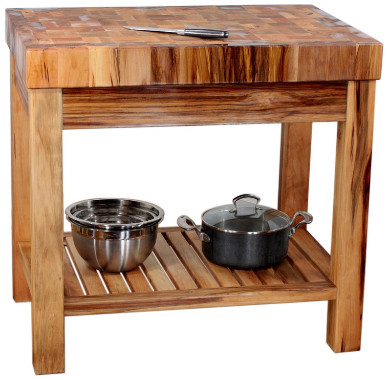May include: A wooden kitchen island with a butcher block countertop and a lower shelf. The island is made of natural wood with a light brown finish. The countertop has a checkered pattern and is made of different types of wood. The lower shelf is made of slats of wood and is empty.