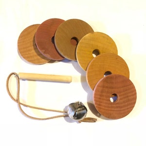 May include: A set of wooden rings with a bell and a leather cord. The rings are different shades of brown and have a hole in the center. The bell is silver and has a small leather loop.