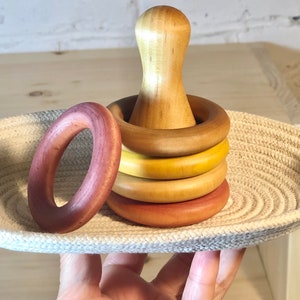 May include: A set of wooden stacking rings in shades of brown, yellow, and orange. The rings are arranged in a small woven basket. The largest ring has a wooden knob in the center.