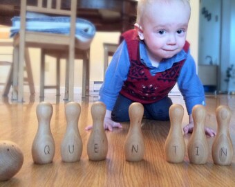 May include: A young child in a red and white sweater is playing with wooden bowling pins that spell out the word "Quentin".