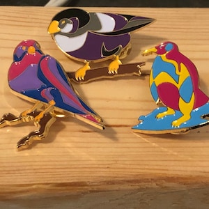 May include: Three enamel pins featuring a purple and blue bird on a branch, a purple and pink bird on a branch, and a yellow, blue, and pink penguin on a blue ice floe.