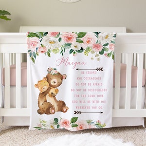 May include: A white baby blanket with a floral design and the name "Maegan" in pink script. The blanket features two cartoon bears and inspirational text. The blanket is draped over a white crib.