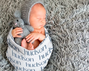 Może przedstawiać: Noworodek w szarym dzianym czapce i owinięty w biały kocyk z nadrukiem imienia "hudson". Dziecko trzyma szarą pluszową myszkę.