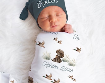 May include: A white swaddle blanket with a repeating pattern of brown dogs holding ducks, green grass, and flying ducks. The blanket has the name "Holden Blake" written on it. A baby is wearing the swaddle and a dark blue knotted hat with the name "Holden" embroidered on it.