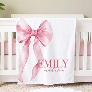 May include: A white baby blanket with a large pink watercolor bow design and the name "Emily Madison" in pink script. The blanket is draped over a white crib with a pink sheet.