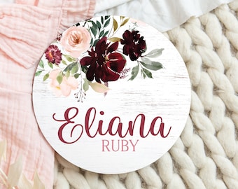 Può includere: Un cartello rotondo in legno con sfondo sbiancato e un motivo floreale in tonalità bordeaux e rosa. Il cartello riporta il nome "Eliana Ruby" scritto in corsivo bordeaux.