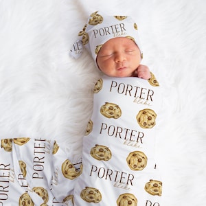 Könnte beinhalten: Eine weiße Wickeldecke mit einem Muster aus Schokoladenkeksen und dem Namen "PORTER blake", der sich wiederholt. Ein Baby ist in die Decke gewickelt und trägt eine passende Mütze mit dem gleichen Muster und Namen.