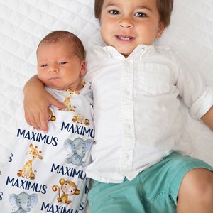 May include: A white swaddle blanket with a blue and white pattern of animals and the name "Maximus" repeated throughout. A baby is wrapped in the blanket and is being held by an older child wearing a white shirt and green shorts.