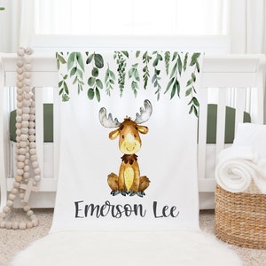 May include: A white baby blanket featuring a watercolour illustration of a moose and green leaves. The blanket is personalised with the name "Emerson Lee".