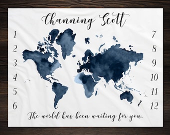 May include: A white baby blanket with a blue watercolor world map and the text "Channing Scott" and "The world has been waiting for you."  The blanket has a number scale from 1 to 12 on the right side.