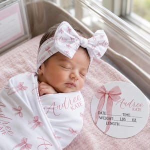 May include: A white swaddle blanket and matching headband featuring a pink bow pattern and the name Audree Kate. A circular white sign displays a pink bow illustration, the name Audree Kate, and blank lines for date, time, weight in lbs and oz, and length.