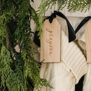 Op de afbeelding: Vier witte gebreide kousen met houten labels die aan een schoorsteenmantel hangen. De labels zijn gepersonaliseerd met de namen "Mommy", "Daddy", "Harper" en "Grayson".