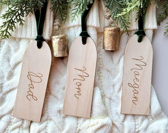 Op de afbeelding: Drie boogvormige houten cadeaulabels met de namen Dad, Mom en Maegan in sierlijk schrift gegraveerd. Elk label hangt aan een donkergroen fluwelen lint naast een rustieke goudkleurige metalen bel, rustend op een witte gebreide kous.