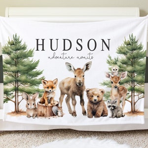 May include: White baby blanket with a woodland theme. It features a moose, fox, rabbit, bear, deer, and raccoon. The blanket has the name "HUDSON" and the text "adventure awaits" printed on it.